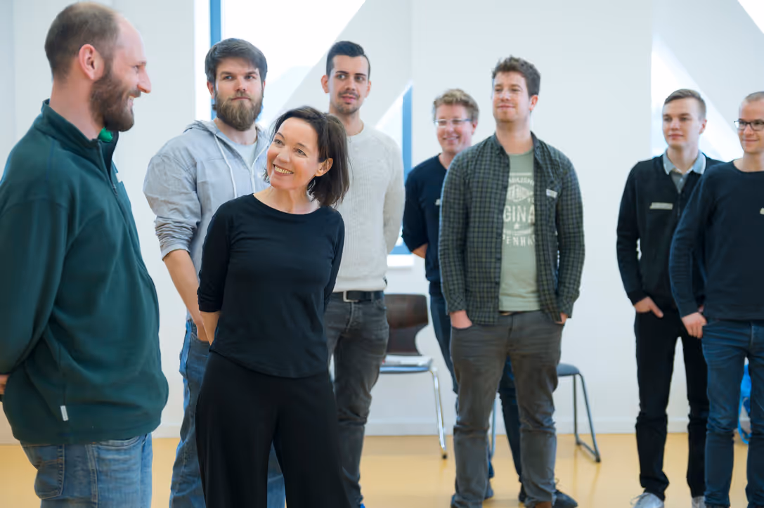 A group of eight adults standing indoors, with a woman in front smiling and engaging with a man on the left.